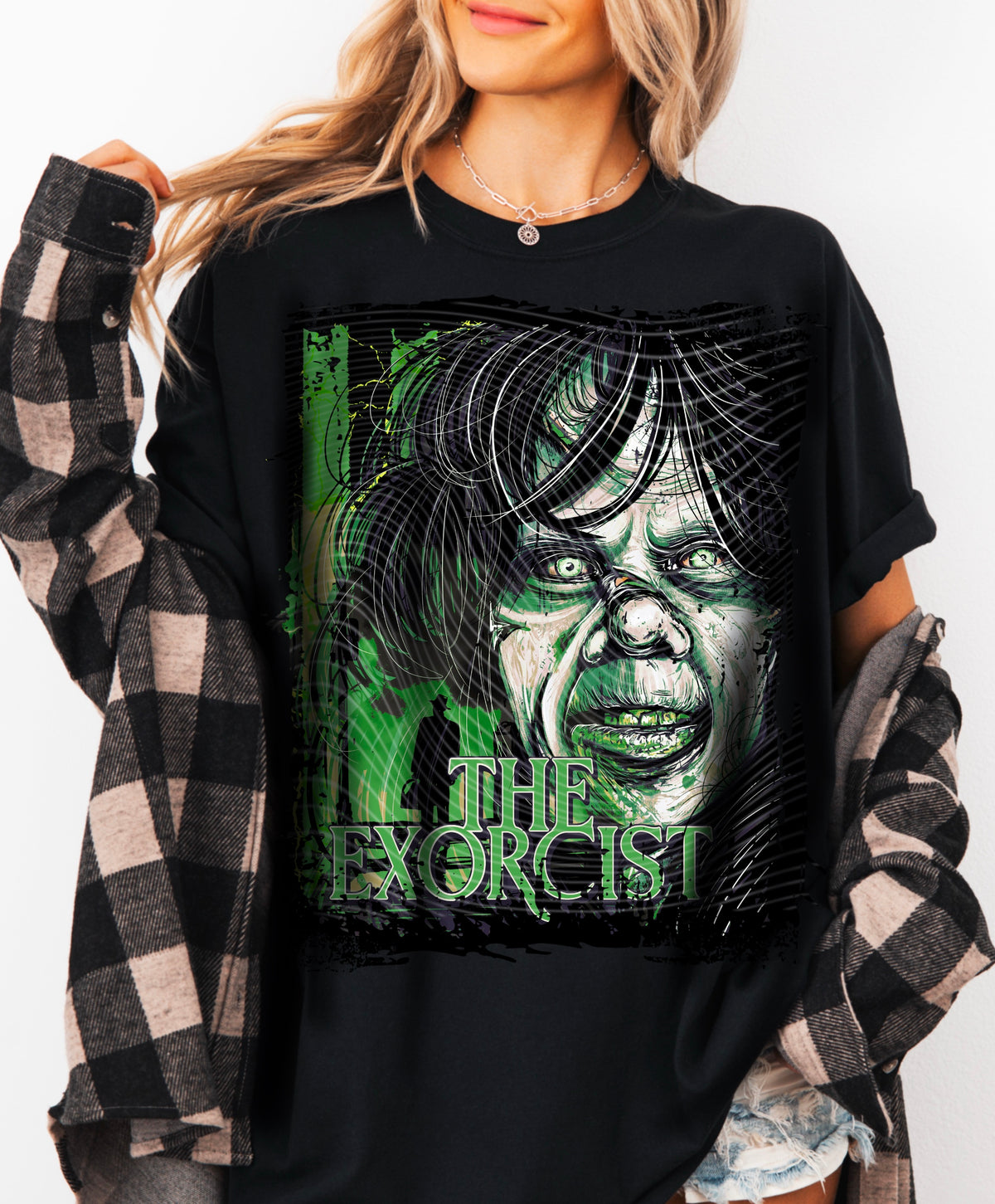 A woman wearing a black oversized graphic T-shirt with a green and gray horror-style face print and the words 'ALL THE EXORCIST'. She has a black-and-beige plaid flannel shirt draped off her shoulders against a plain white background.