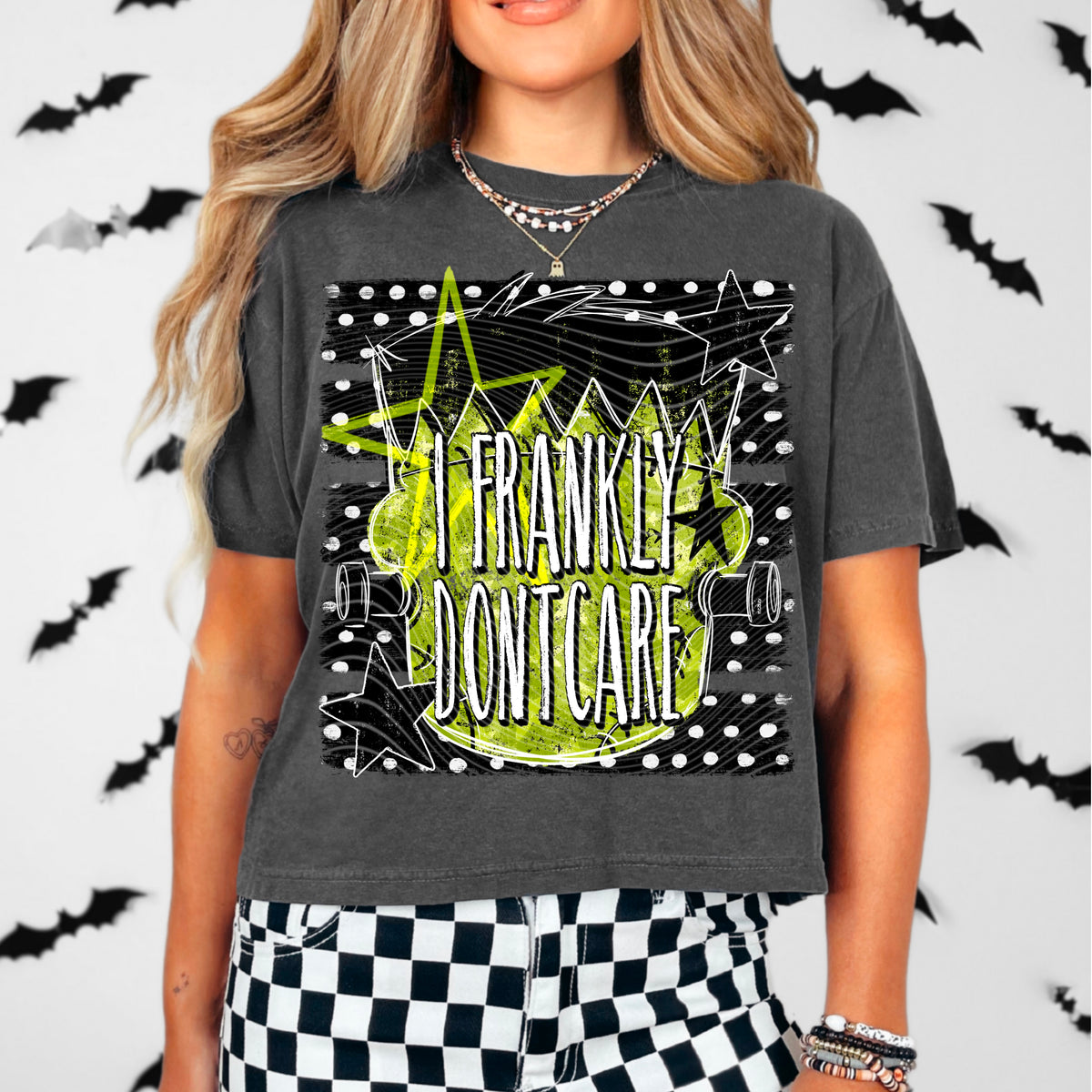 A woman wearing a dark charcoal gray graphic t-shirt with a lime green and white 'I FRANKLY DONT CARE' design, paired with black and white checkered pants against a white background decorated with black bat silhouettes.