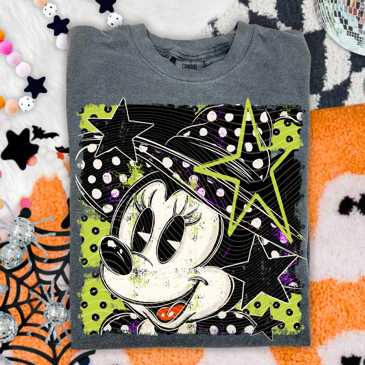 A folded gray t-shirt featuring a graphic of Minnie Mouse in a witch hat with neon green stars and black polka-dot details, styled on a white and orange patterned blanket with Halloween-themed props.