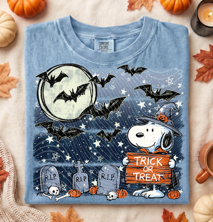 A light blue graphic T-shirt laid flat on a neutral fabric surface with autumn props. The shirt features a Halloween scene with bats, a full moon, tombstones, pumpkins, and Snoopy holding a 'Trick or Treat' sign.