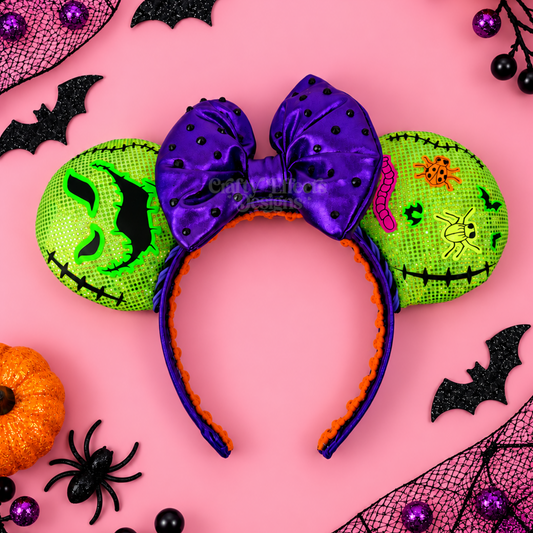 A Halloween-themed mouse ears headband featuring a large purple bow, one green pumpkin-shaped ear with a stitched face, and one decorated with a spider and moon motif, set against a pink background with Halloween decorations.