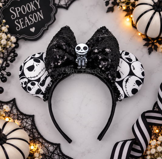 A gothic Disney Minnie Mouse ears headband styled for Halloween, featuring a large black sequined bow with a Jack Skellington charm, black-and-white patterned ears, and a black headband, displayed on a marble surface with spooky props.