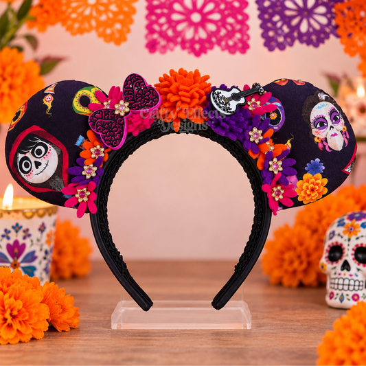 A Day of the Dead themed mouse ears headband on a clear acrylic stand, decorated with colorful fabric flowers, sugar skull patterns, and a purple glittery bow, set against a festive background with papel picado and marigolds.
