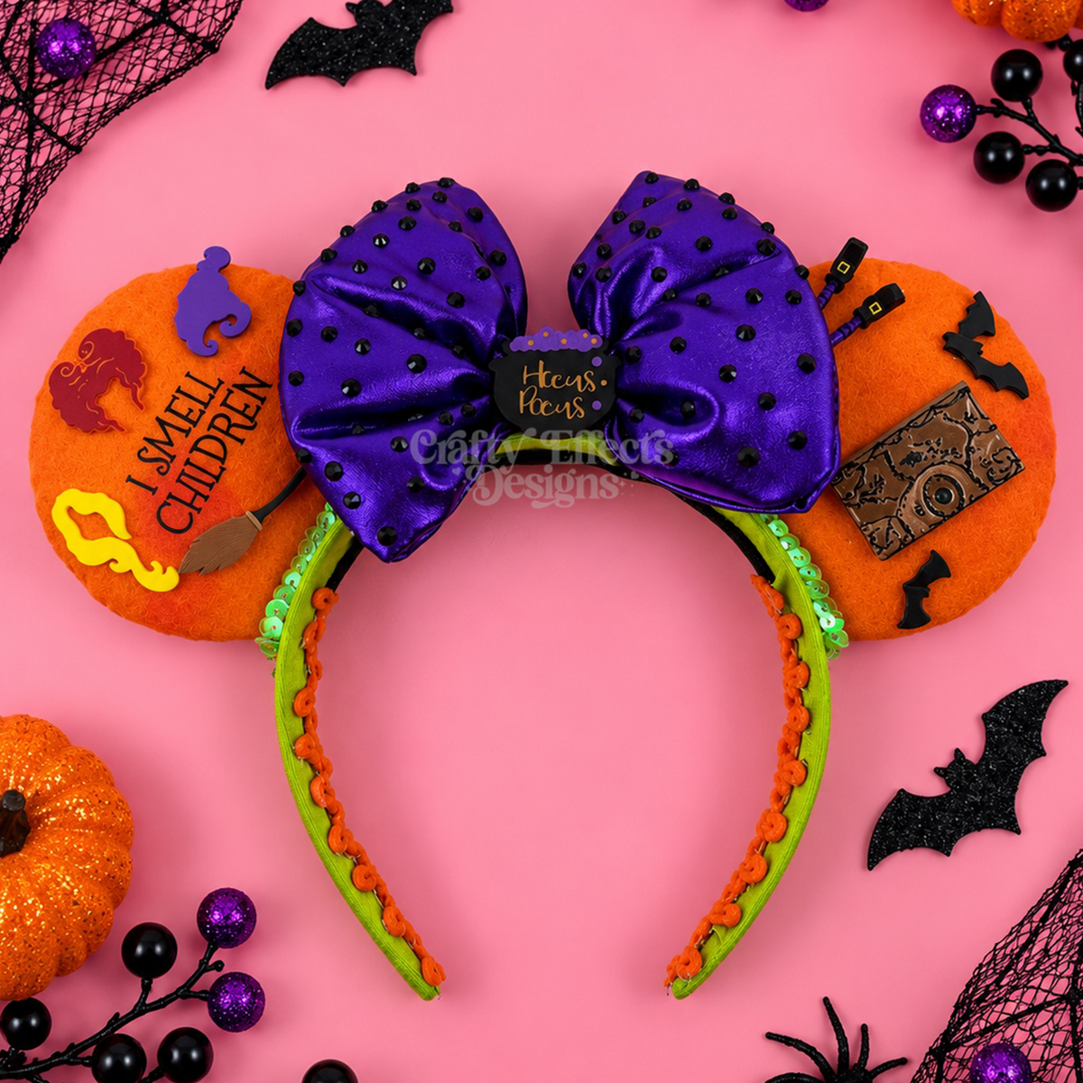 Halloween-themed mouse ears headband featuring a large purple bow with black polka dots, orange ears with witch hats, broom, and cauldron embellishments, and a green-and-orange spiked band. The headband is styled on a pink background with Halloween decorations including bats, black lace, purple beads, and orange pumpkins.